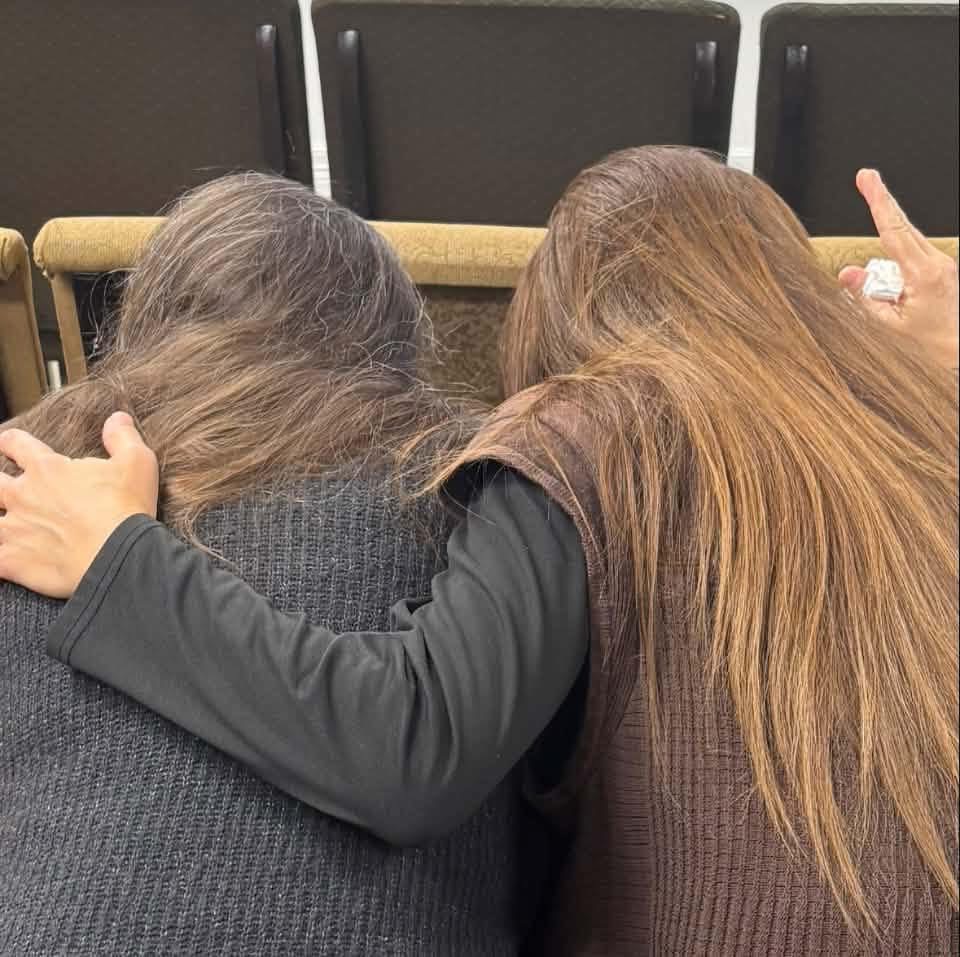 Two women sitting side by side, one with her arm around the other, heads bowed together